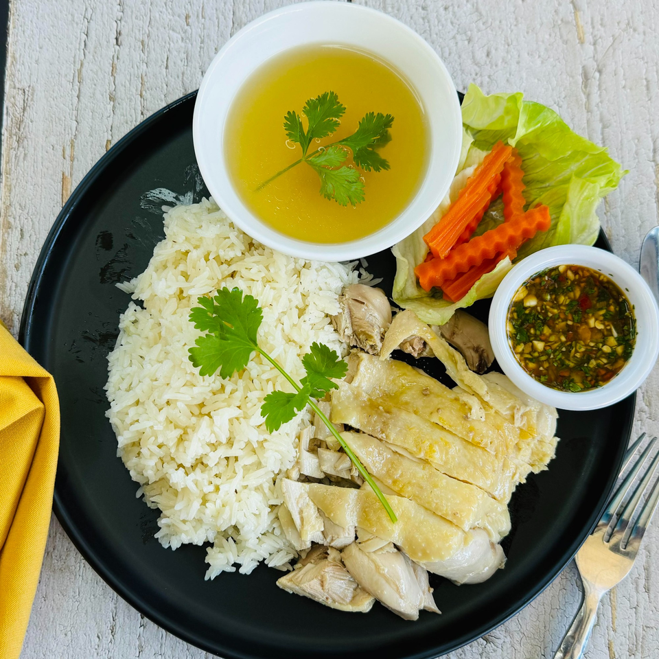 Hainanese chicken rice with sliced chicken, rice, broth, sauce, and vegetables on a black plate.