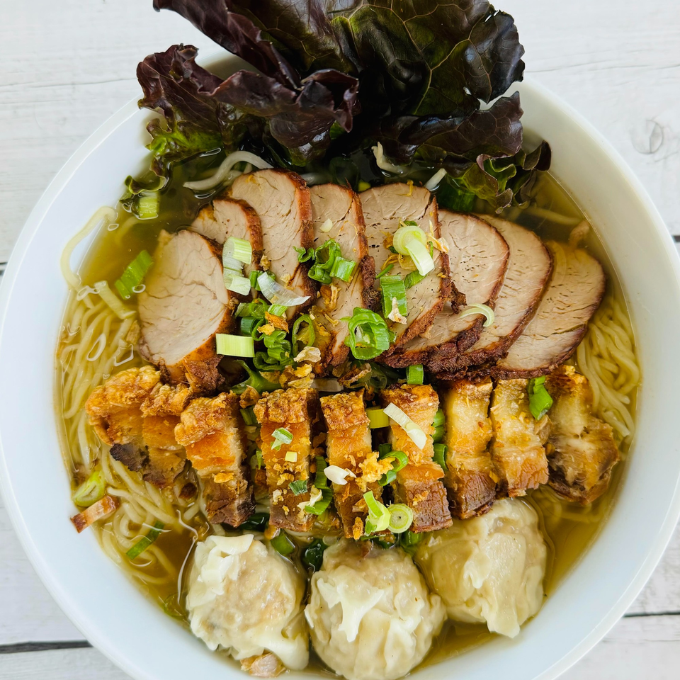 A bowl of Thai noodle soup with sliced pork, crispy pork belly, wontons, and green onions.