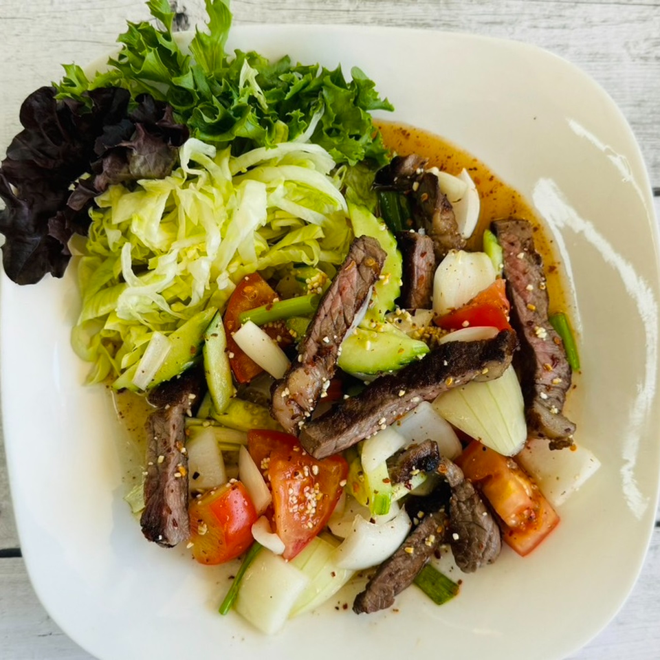 Thai beef salad with sliced steak, tomatoes, onions, cucumbers, and mixed greens on a white plate.