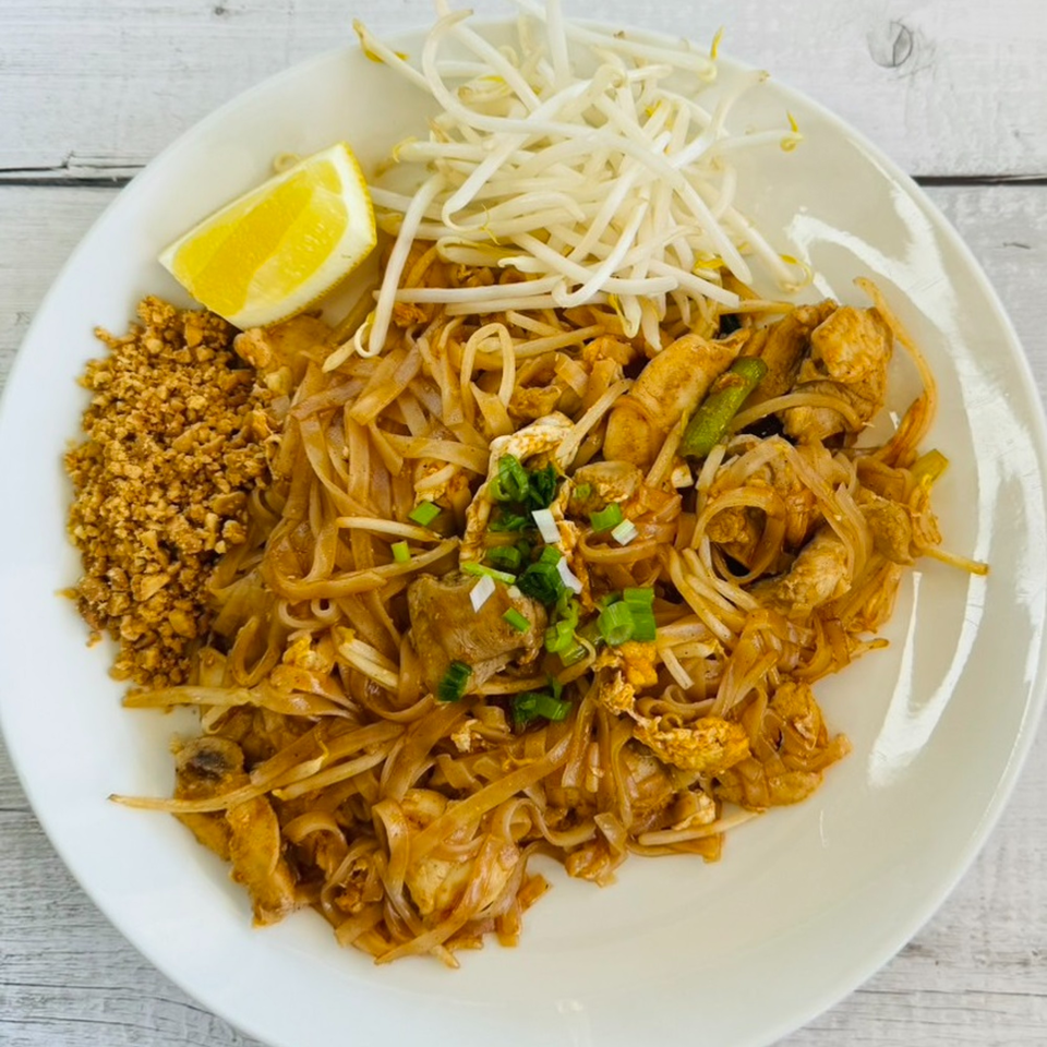 A white plate of Pad Thai with chicken, bean sprouts, crushed peanuts, and a lemon wedge.