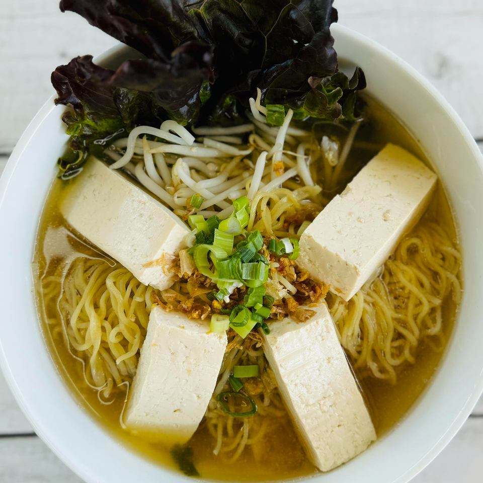 Thai noodle soup with tofu, bean sprouts, green onions, and dark leafy greens in a white bowl.